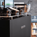 A black Ascaso espresso machine with a silver logo, shown with several glossy black coffee cups in a metal rack and a glass bean hopper filled with coffee beans. The background is a softly blurred cafe interior.