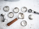 Flat-lay of stainless steel espresso accessories including wooden portafilter, tamper, and filter baskets